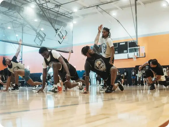 A group of basketball players stretching on an indoor court. They are wearing jerseys and are in various stretching positions, with one person standing and instructing them. The court has a basketball hoop and a scoreboard in the background.